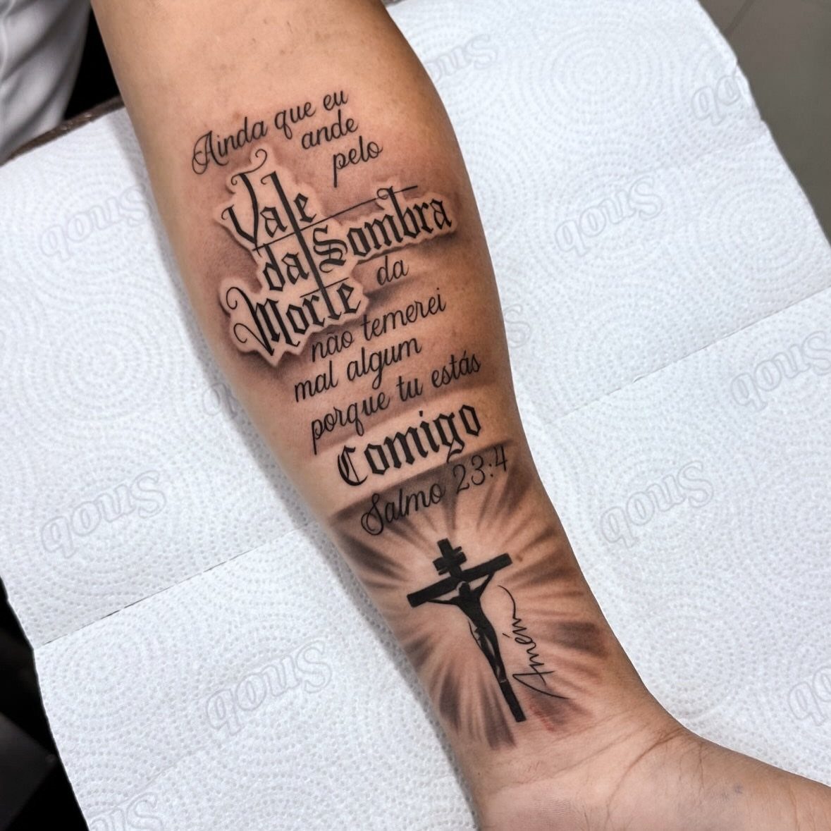 A forearm tattoo featuring large cursive Portuguese script and a small shaded crucifix.