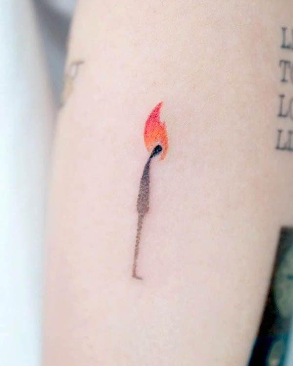 A small minimalist tattoo of a lit match with an orange flame on skin.