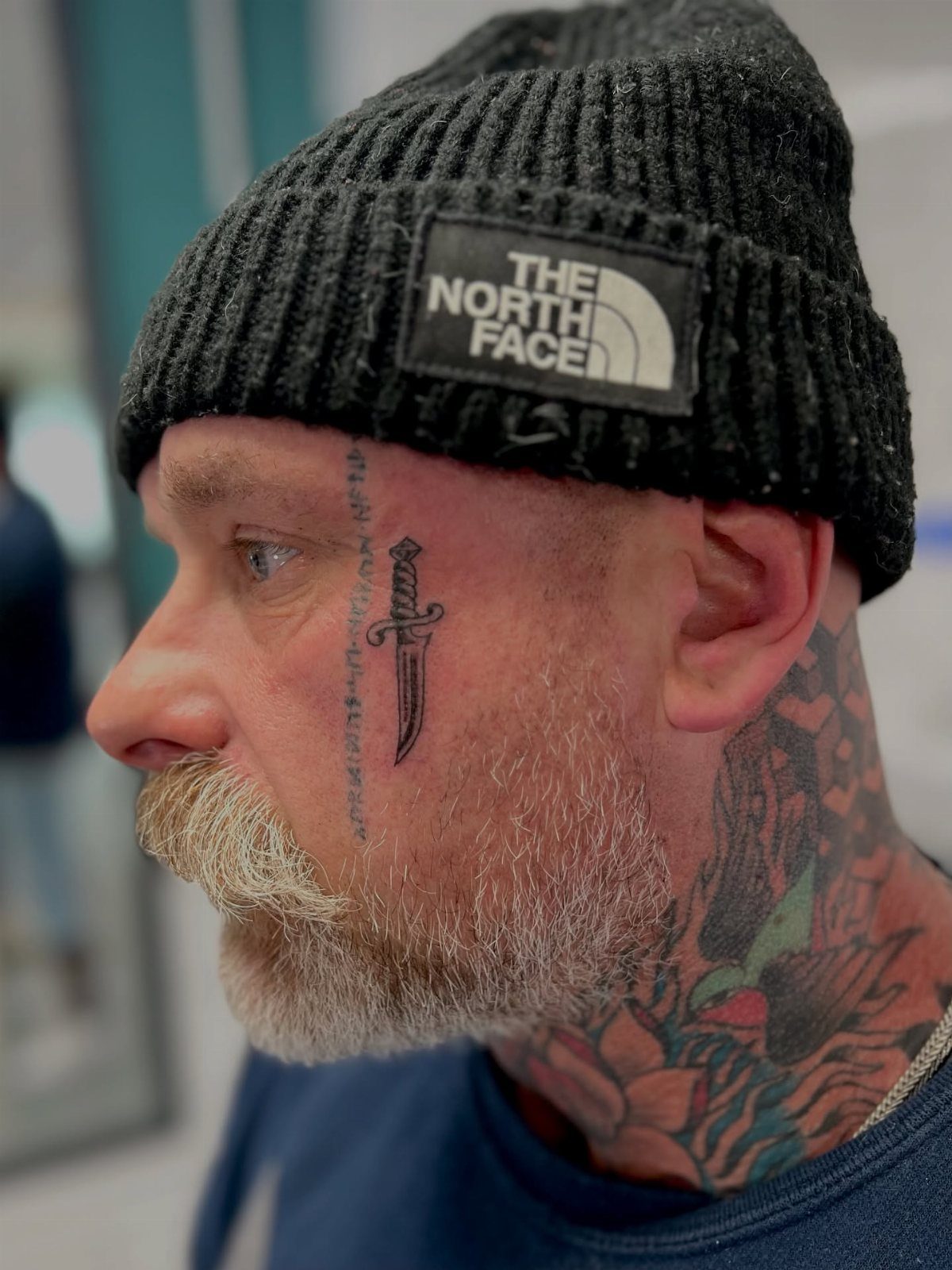 Man with a grey beard and black beanie featuring a dagger tattoo on his cheek.