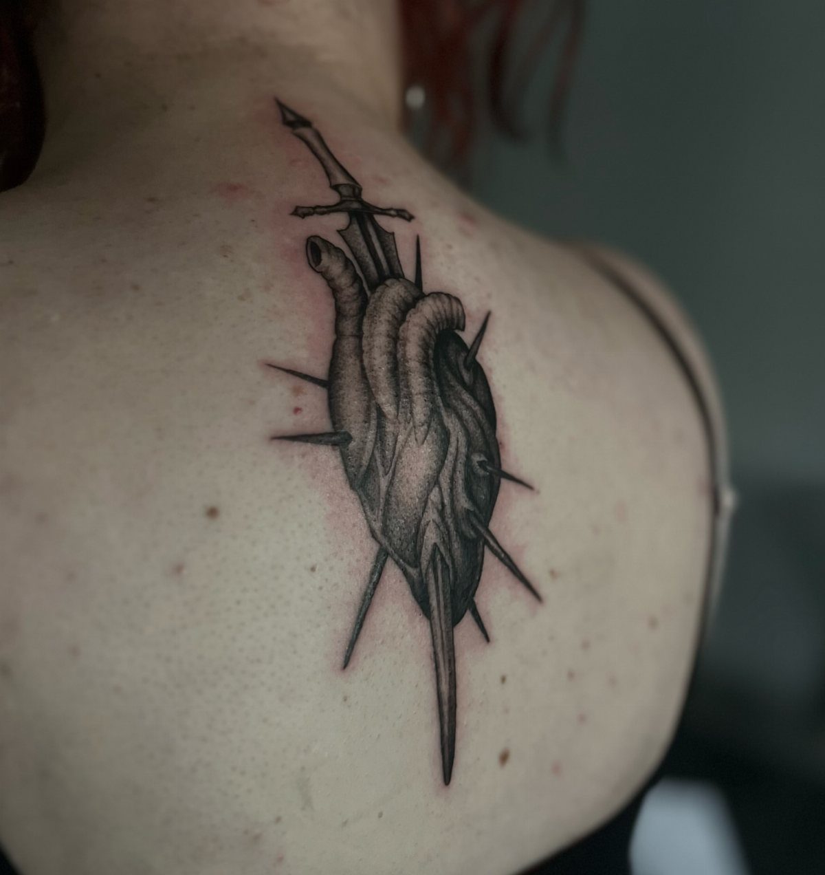 Anatomical heart tattoo pierced by a dagger on a person's upper back.