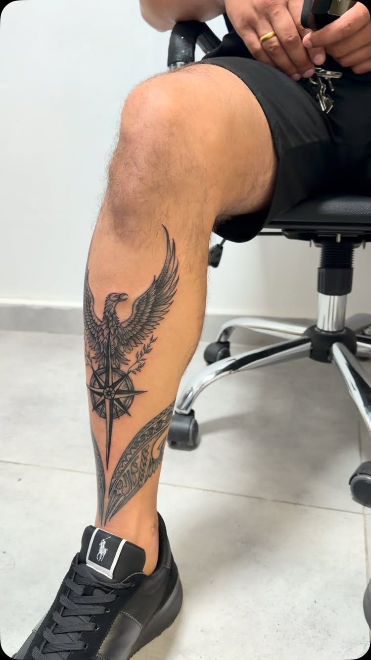 A large black ink eagle and compass tattoo on a man's lower leg.