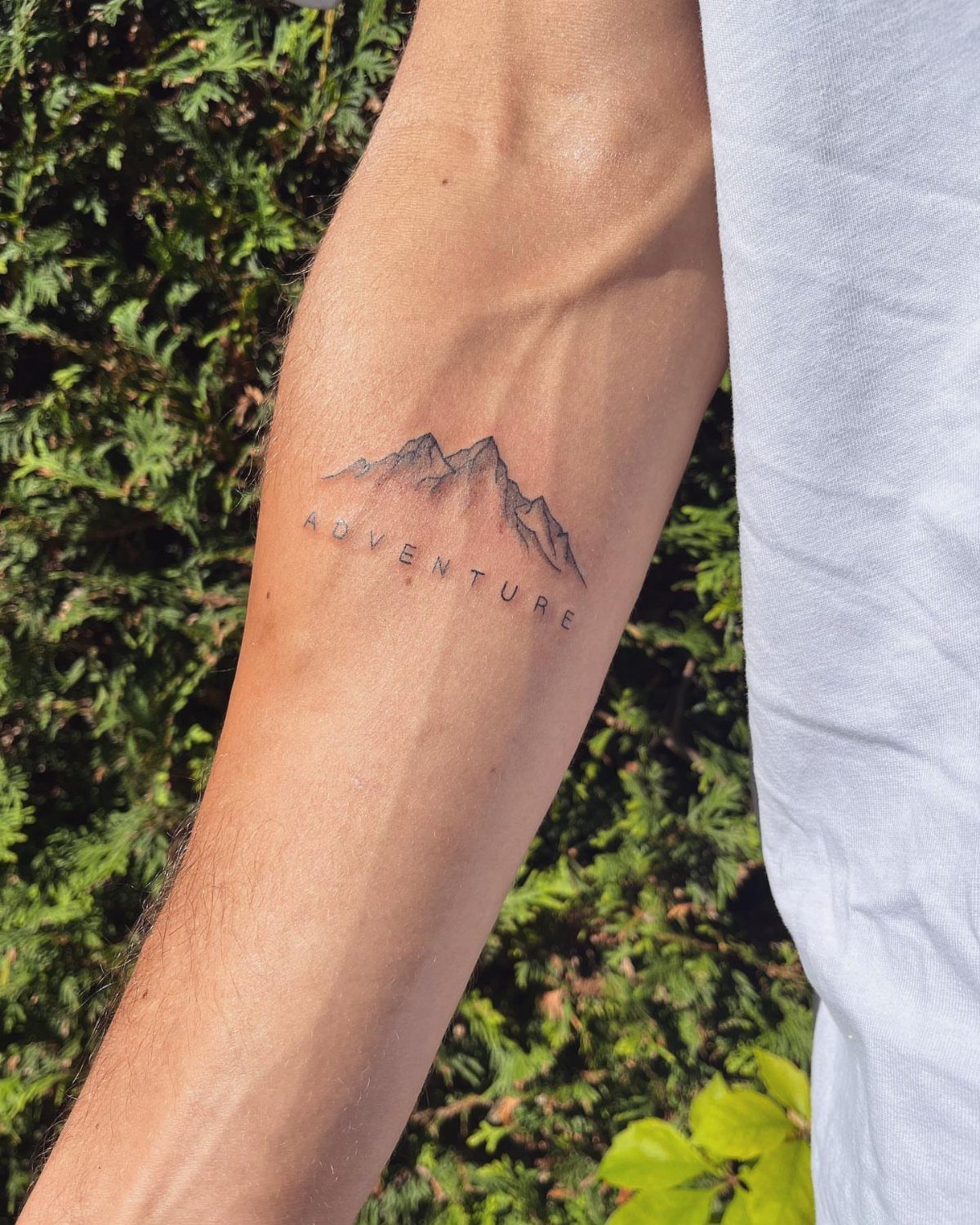 A fine-line mountain range tattoo with the word adventure on a person's forearm.