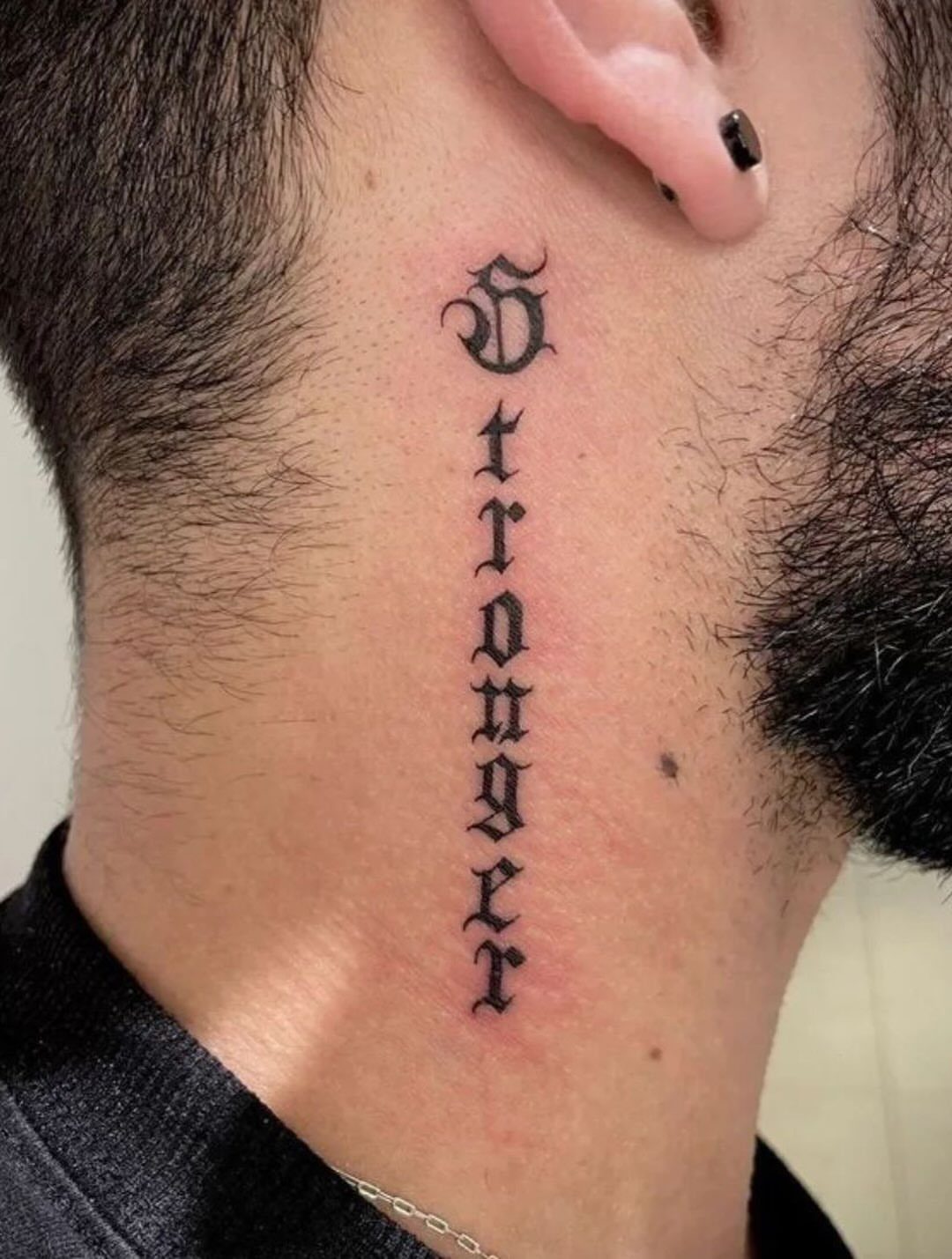 A vertical black ink gothic font tattoo reading 'Stronger' on a man's neck.